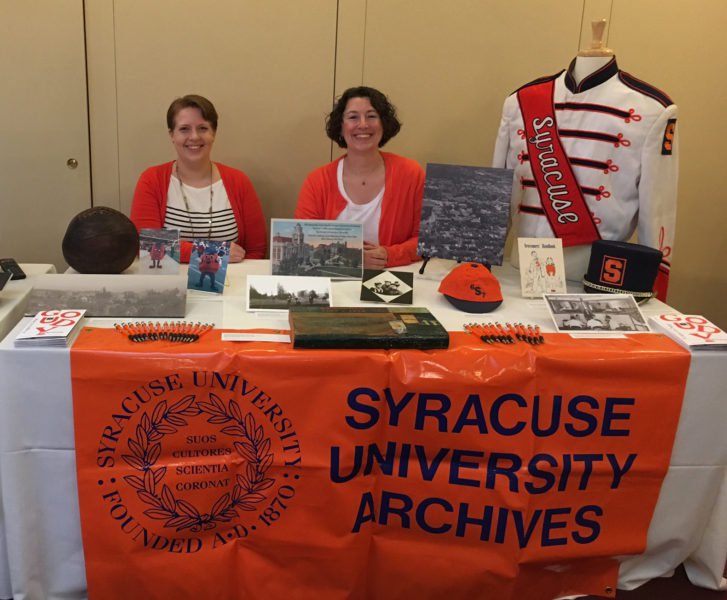 Two women wearing orange cardigans sitting behind Syracuse University Archives table, with memorabilia including band uniform on table