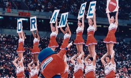 SU cheerleaders on basketball court holding up letters that read Orange and Otto the Orange mascot standing behind them; fans sitting in stands