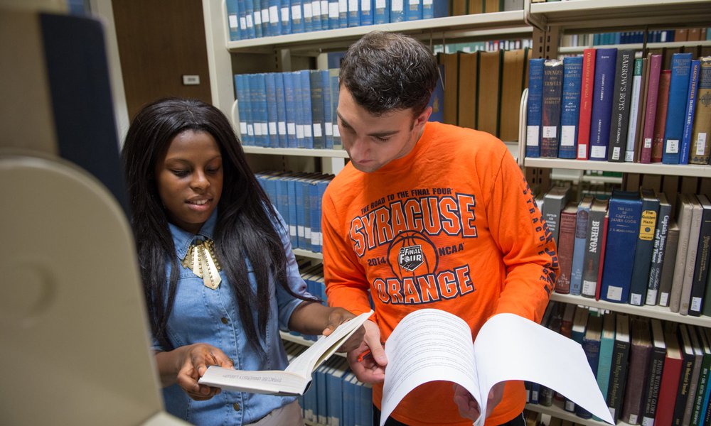 Patrons in the library; 2 students, one Black woman with long hair and one white male wearing orange shirt, looking at materials next to book shelves