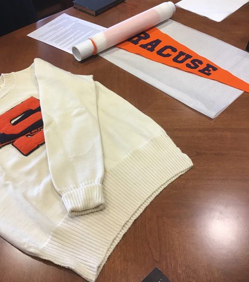 A white sweatshirt with an embroidered orange letter ‘S’ and an orange textile stating “Syracuse” on top of a brown table.