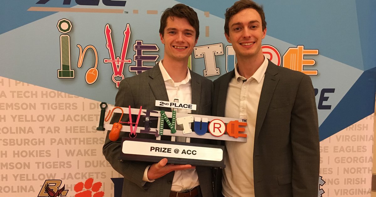 Two students wearing suits and holding ACC Inventure Prize trophy in front of matching step and repeat banner