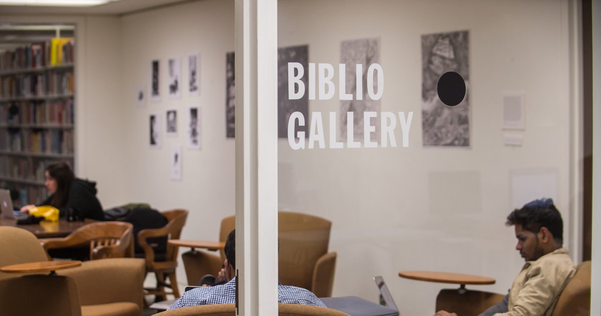 Several students sitting inside glass walls of the Biblio Gallery with artwork on wall and bookshelf on other side