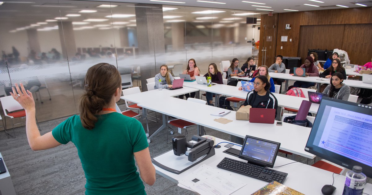 Instructor leading a classroom of students sitting at long white tables