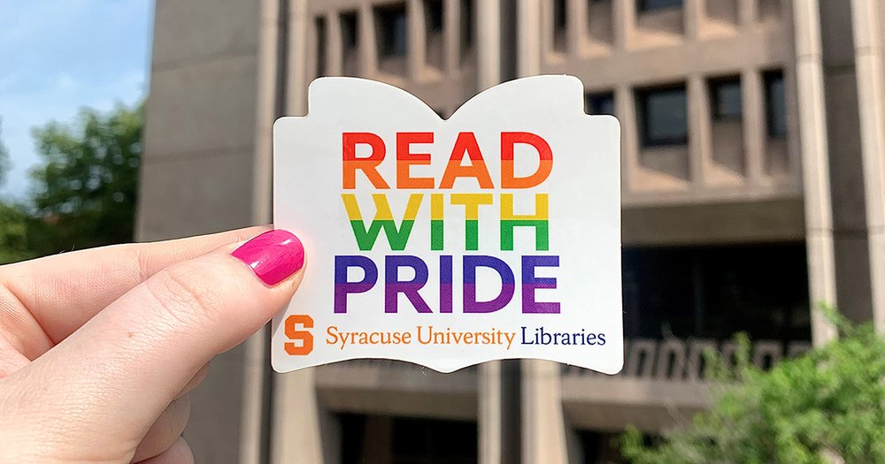 Book-shaped white sticker with rainbow text that says Read With Pride and Syracuse University Libraries logo being held up in front of Bird Library