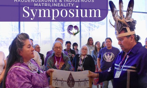 woman holding wampum belt on left, man wearing feather headdress on right, people in background with symposium title at top