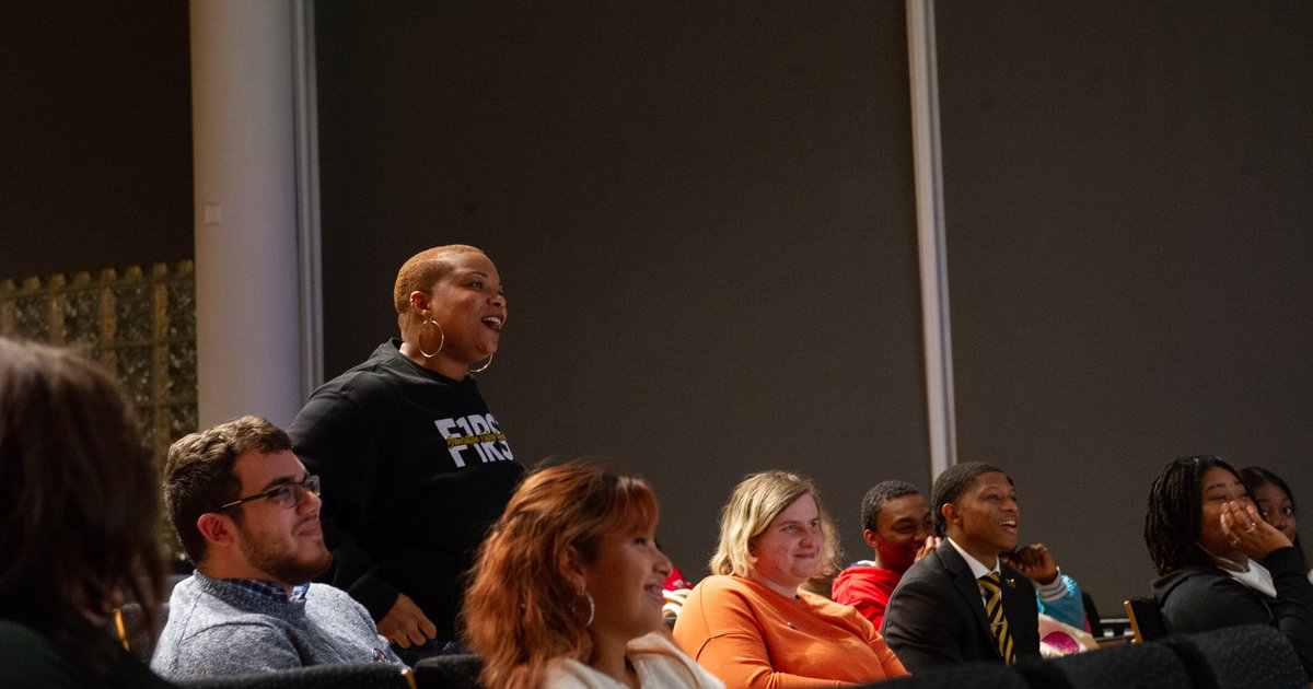 Black woman wearing black sweatshirt standing amid group of people sitting in audience