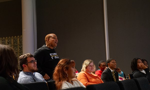 Black woman wearing black sweatshirt standing amid group of people sitting in audience