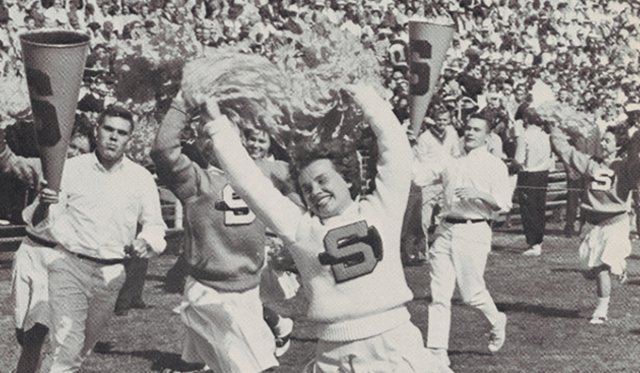 Archival black and white photo of female Syracuse University cheerleader running with pom poms in Block S sweater with male cheerleaders running behind with megaphones