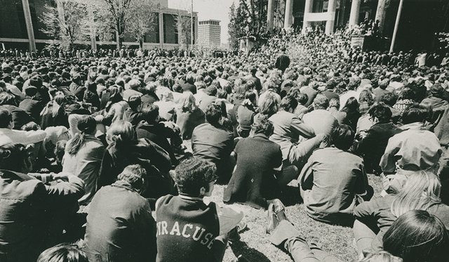 Black and white archival image of hundreds of students seated on the quad in front of Hendricks Chapel at Syracuse University with backs facing the camera, circa 1970