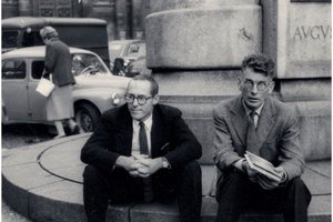b&w photo of two men sitting on curb on side of building