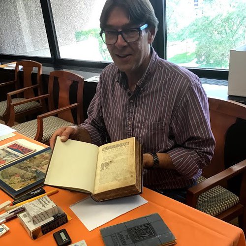 Man with glasses wearing a flannel shirt holding open a book while seated in front of a window and behind an orange table with other books and paper items.