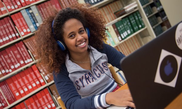 Student wearing over the ear headphones and using a laptop in front of a large book shelf