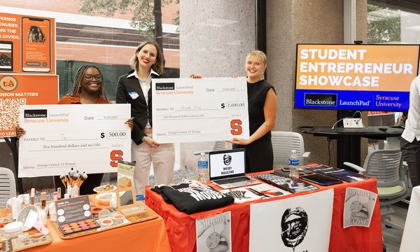 3 students holding up giant checks behind display tables