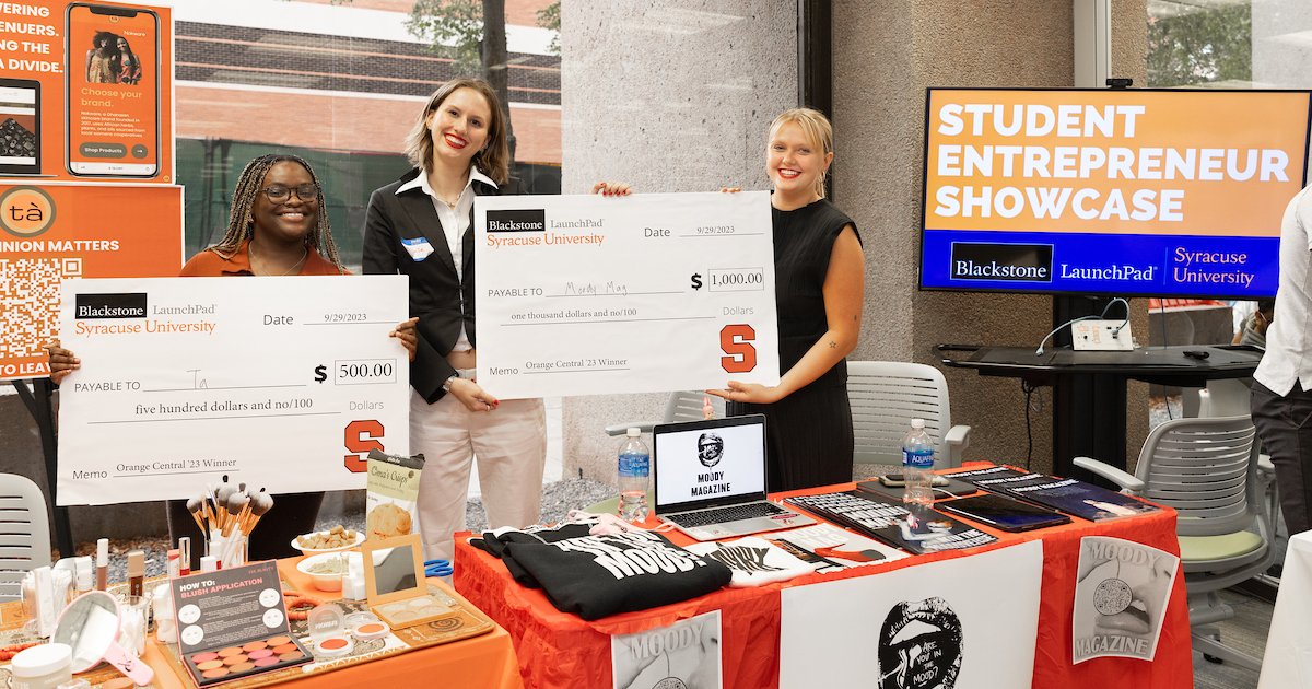 3 students holding up giant checks behind display tables