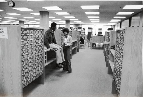 Students talking in long card catalog aisles in Bird Library. Syracuse University Photograph Collection