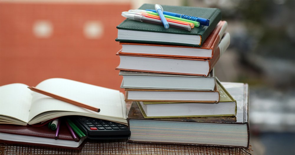 Stack of textbooks with pens on top next to an open notebook with a pen and a calculator