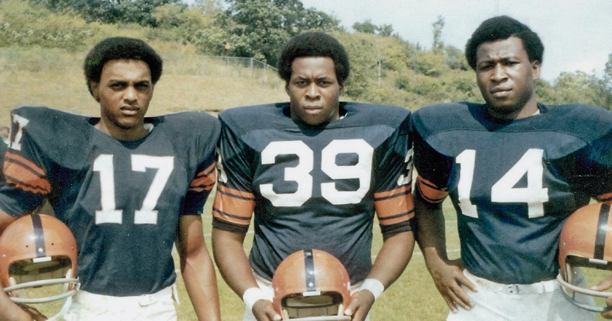 Three Black Syracuse football players in dark blue jerseys, numbers 17, 39 and 14, holding their helmets, members of the Syracuse 8