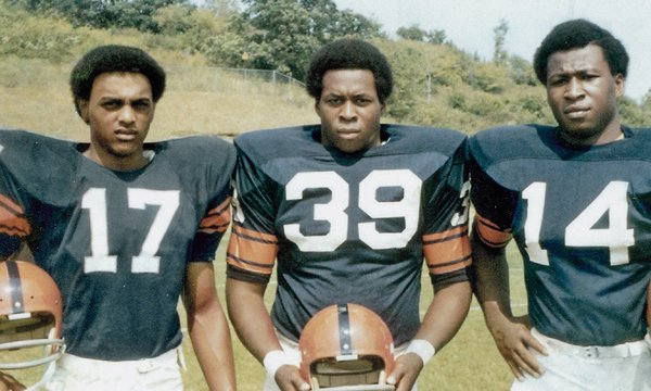 Three Black Syracuse football players in dark blue jerseys, numbers 17, 39 and 14, holding their helmets, members of the Syracuse 8