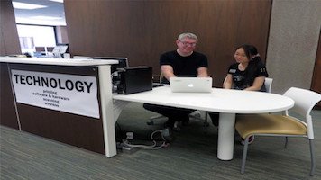 man sitting next to woman in front of laptop. Sign in front of desk reads "Technology"