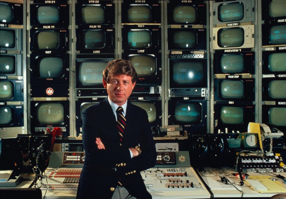 Ted Koppel standing in front of dozens small television screens