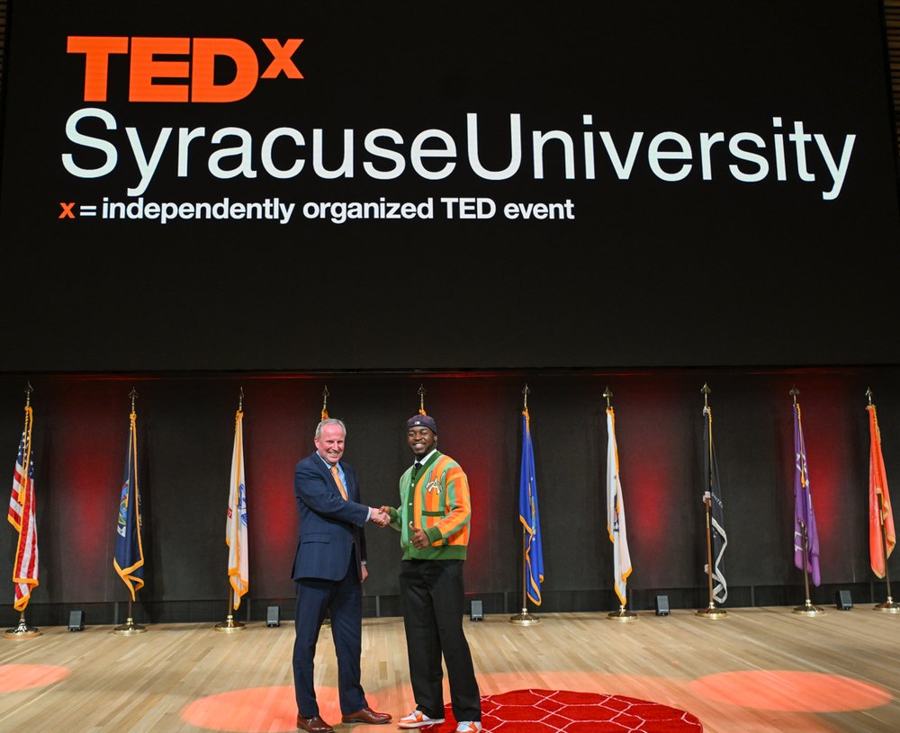 two people shaking hands on stage with sign behind them that reads TEDx Syracuse University