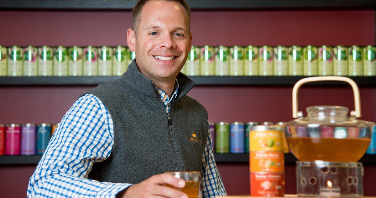 Todd Rubin in a gray vest and blue checkered shirt sitting with tea pot and can of tea in front of a wall lined with tea cans