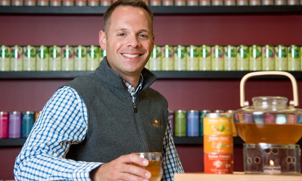 Todd Rubin in a gray vest and blue checkered shirt sitting with tea pot and can of tea in front of a wall lined with tea cans