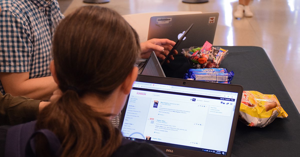 Back of head of a student searching the Syracuse University Libraries catalog on a laptop with snacks on front of the table