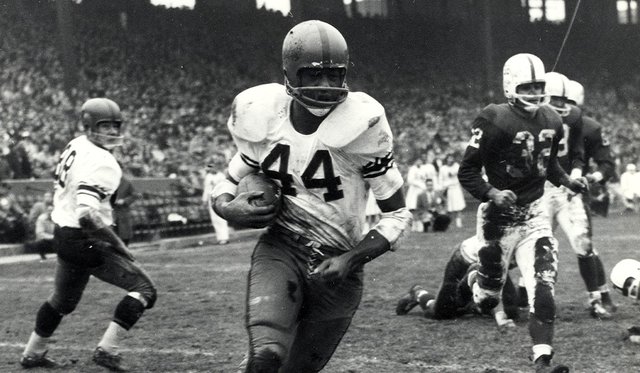 Black and white archival image of a Syracuse University football player in a white #44 jersey and helmet carrying the ball down the field with other players behind him