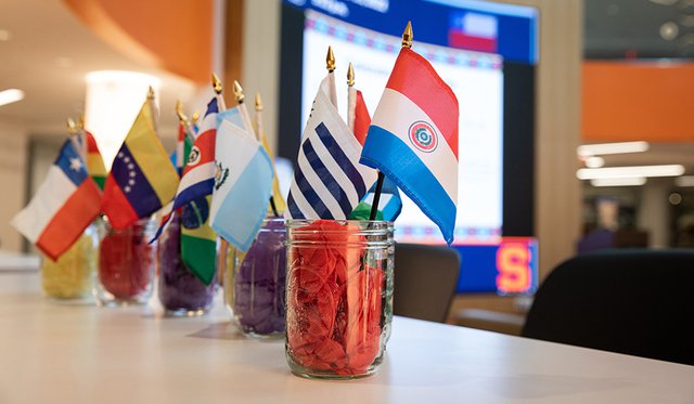 Row of glass jars on a table in front of a large digital screen filled with colorful tissue paper and Latin American country flags