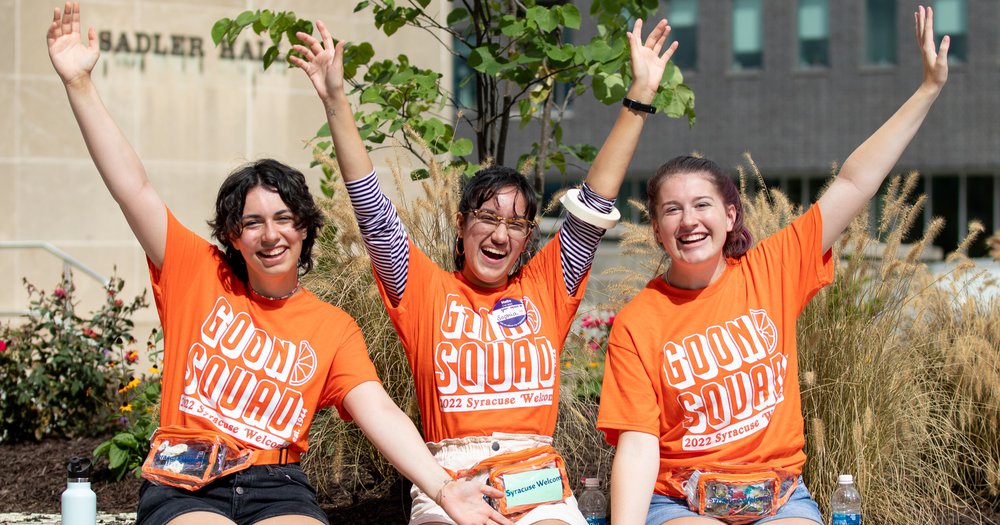 three students sitting side by side wearing orange Goon Squad 2022 Welcome t-shirts and raising their hands in the air