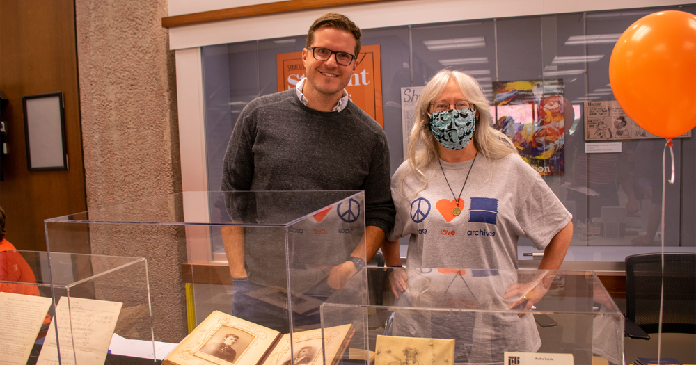 person wearing gray sweater, another person with long hair wearing face mask, standing behind table with materials under glass display and in front of glass display case