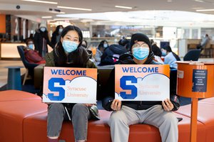 two students sitting side by side wearing masks and holding sign that says Welcome to Syracuse University