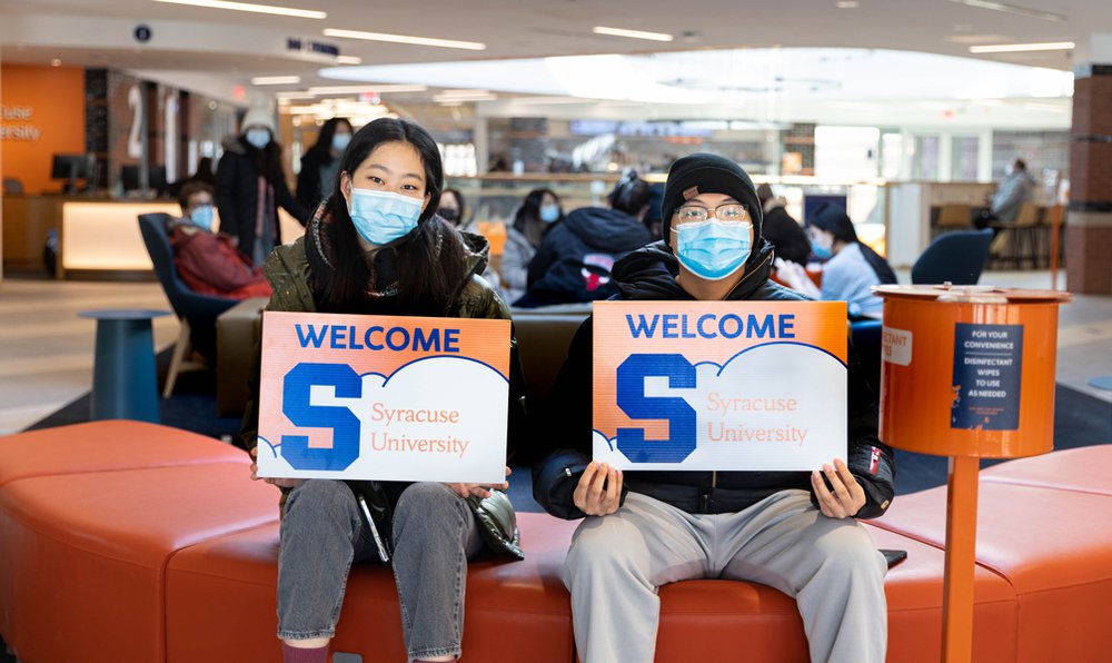 two students sitting side by side wearing masks and holding sign that says Welcome to Syracuse University