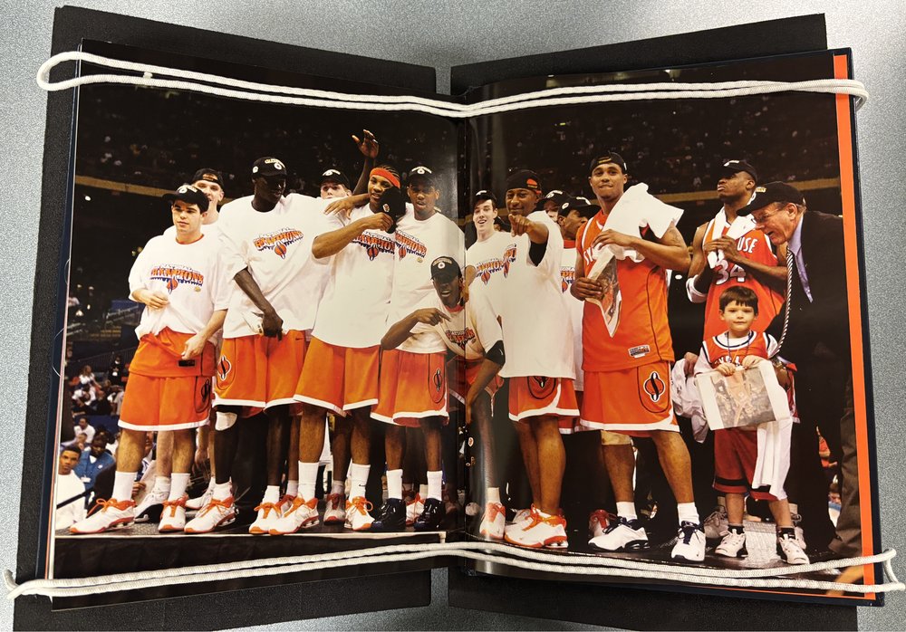The Syracuse University Men’s Basketball team just after their 2003 NCAA Championship win. They wear shirts that read “National Champions” and are celebrating together. Carmelo Anthony is third from the left in the front row.