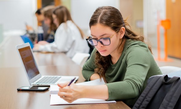 Student writing on a notebook with a laptop on a desk