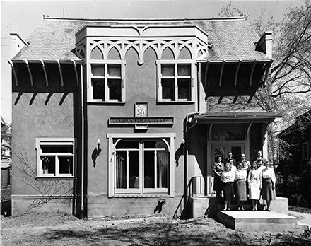 cottage house with ornamentation above window and group of women posing on porch