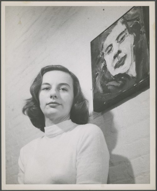 Black and white portrait photograph of a woman in a white sweater in front of a wall with a painting of her self-portrait.