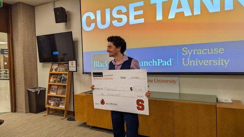 man holding large check in front of digital sign that reads &#x27;Cuse Tank