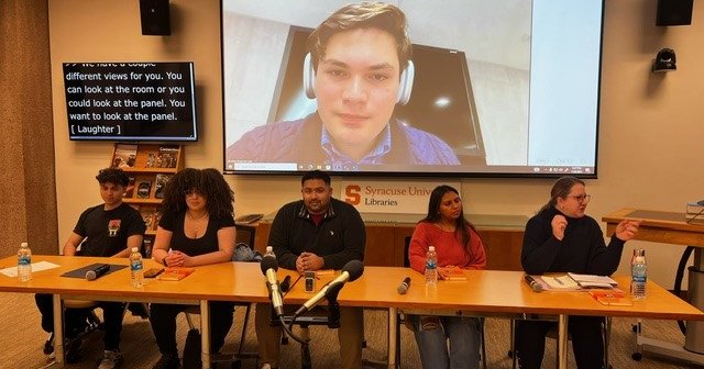 five people sitting behind table in Bird Library with one person on screen behind table