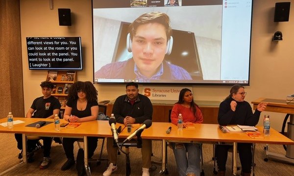 five people sitting behind table in Bird Library with one person on screen behind table