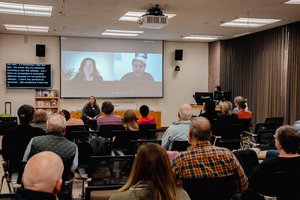 person sitting in front of room with two people on screen behind her, while audience members listen