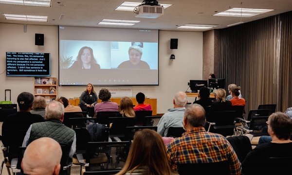 person sitting in front of room with two people on screen behind her, while audience members listen