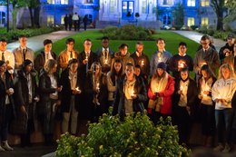 students standing in front of Remembrance Wall outside hall of languages at dusk holding candles