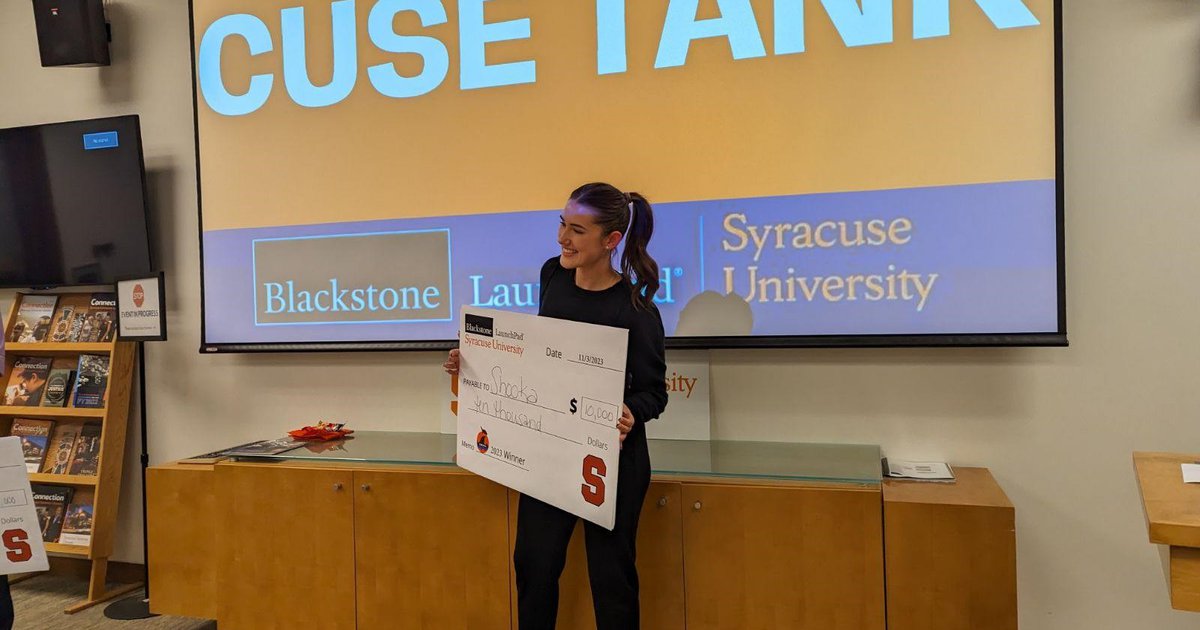 woman holding large check in front of digital sign that reads Cuse Tank
