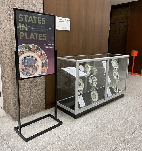 small poster sign that reads "States in Plates, Indiana to Michigan" next to 3-tier glass display case with souvenir plates and descriptions