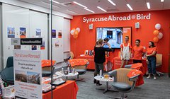 room with bright orange wall that reads Syracuse Abroad and balloons, chairs and people standing