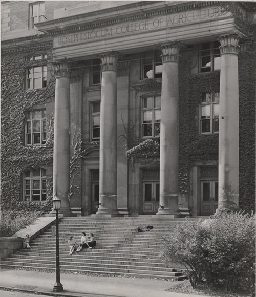 large building with four columns, Joseph Slocum College of Agriculture etched into the stone of the building; several women sitting on steps in front of buildng